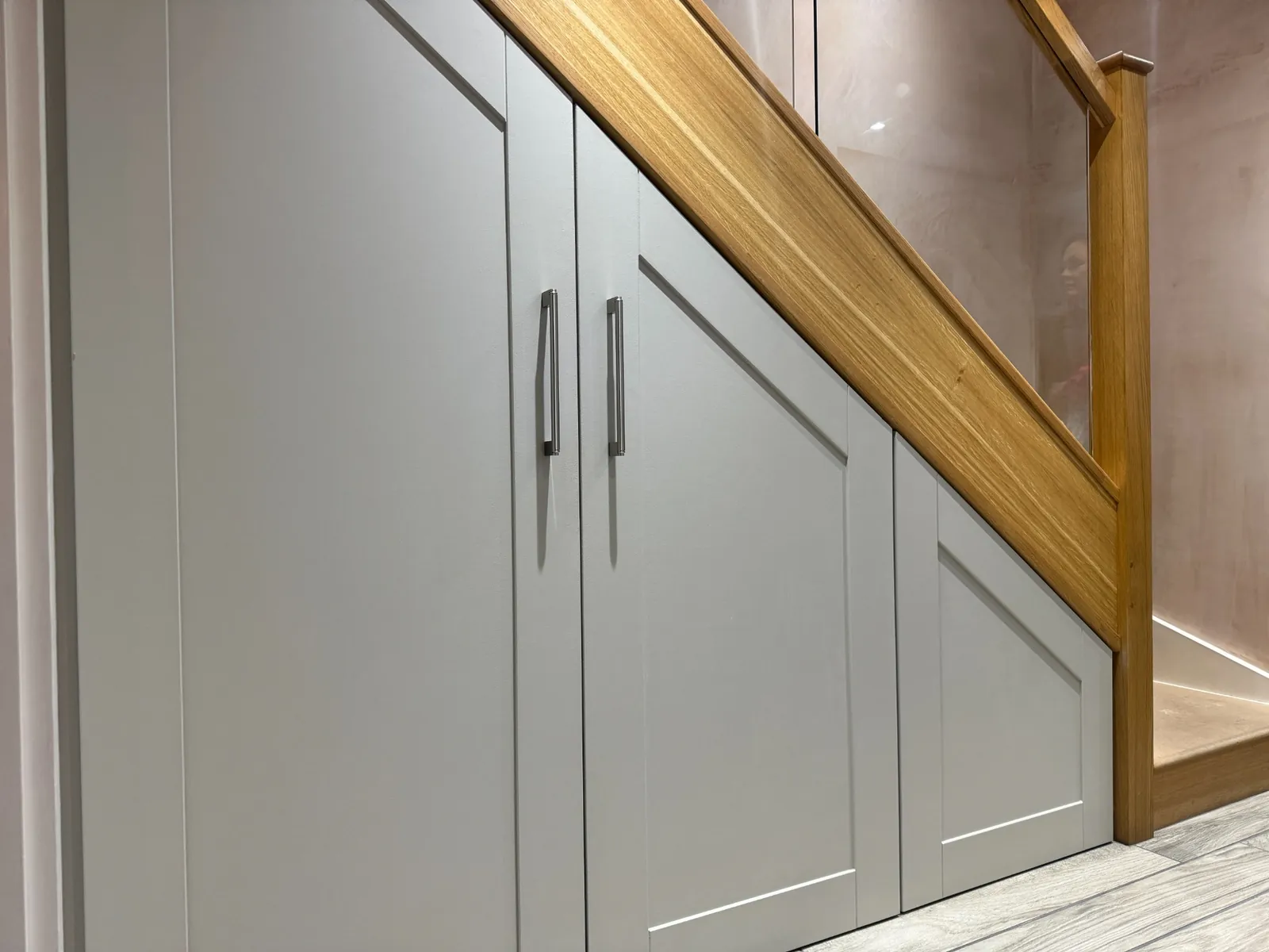 Fitted understairs storage cupboards with shaker doors beneath an oak and glass staircase in a Cheshire home.